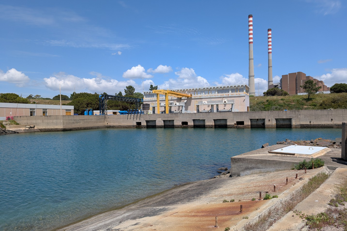 Start_Campus_Portugal_sea_water Industrieel gebouw met twee hoge schoorstenen naast een waterpartij, omgeven door betonnen constructies en een gedeeltelijk bewolkte lucht.