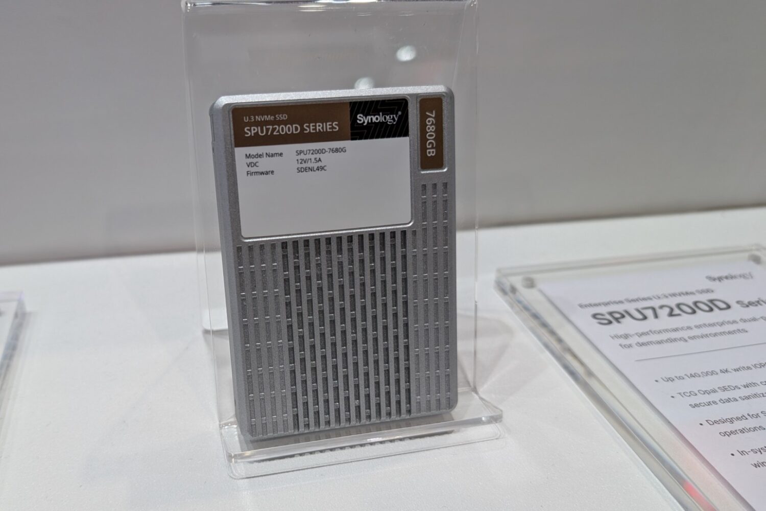 Een Synology SPU7200-serie 7,68TB solid-state drive wordt rechtop gepresenteerd in een doorzichtige plastic standaard op een wit oppervlak, naast de Synology PAS7700 voor naadloze opslagoplossingen met hoge capaciteit.
