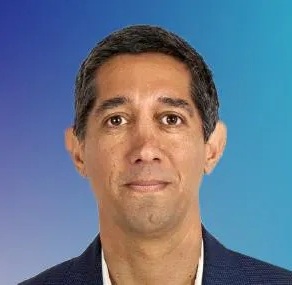 Een man met kort, donker haar, gekleed in een donker colbert en een wit overhemd, wordt afgebeeld tegen een blauwe achtergrond.
