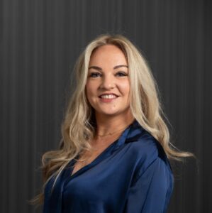 Een vrouw met lang blond haar en een blauwe satijnen blouse lacht naar de camera tegen een donkere, verticaal gestreepte achtergrond.