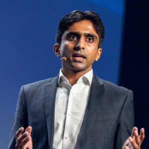 Een man in pak spreekt op het podium met een headset-microfoon en gebaart met zijn handen tegen een blauwe achtergrond.
