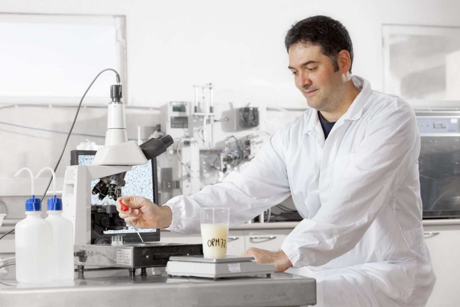 Hendrix Genetics (Large) Een wetenschapper in een witte labjas onderzoekt een monster onder een microscoop in een laboratorium, met labapparatuur en een bekerglas met het label "ORM" zichtbaar op de tafel.