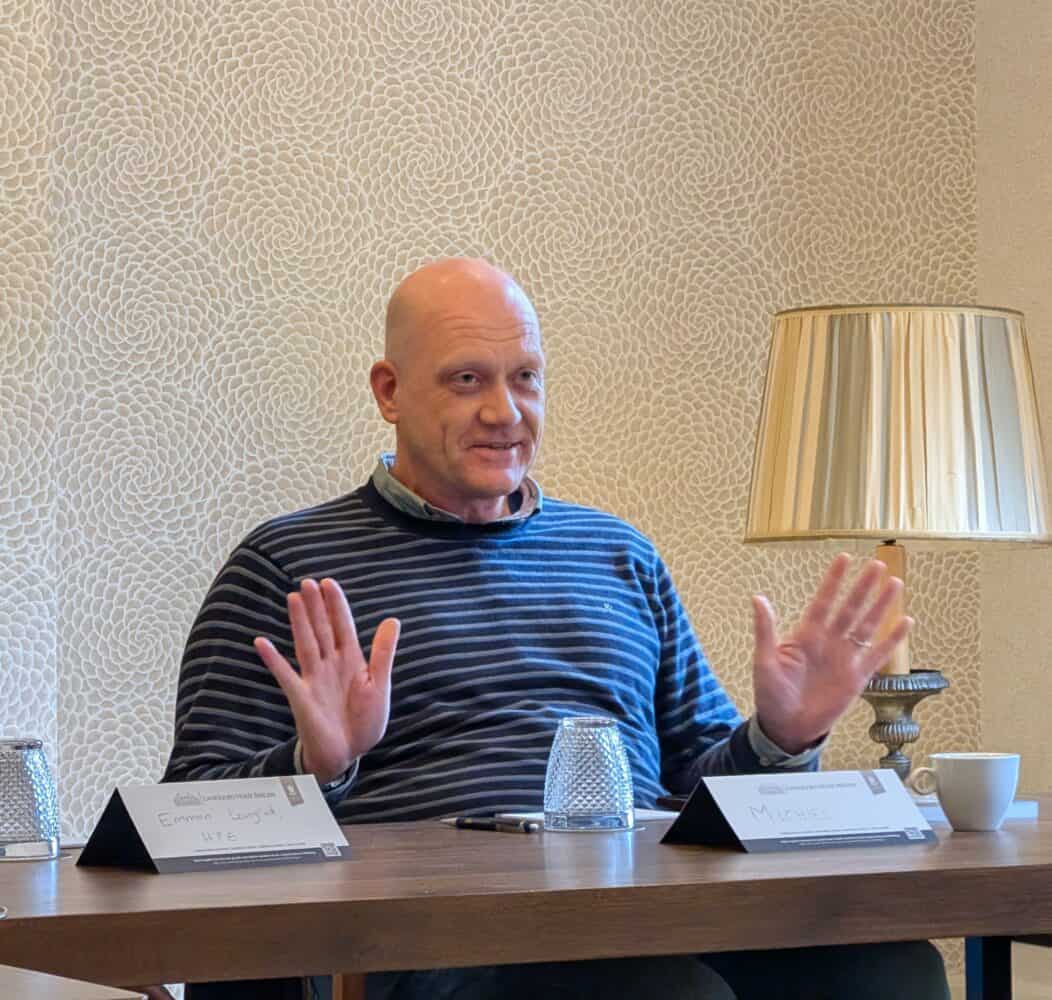 Een kale man in een gestreepte trui zit aan een tafel met een lamp, twee naamplaatjes en glaswerk en gebaart met beide handen omhoog.