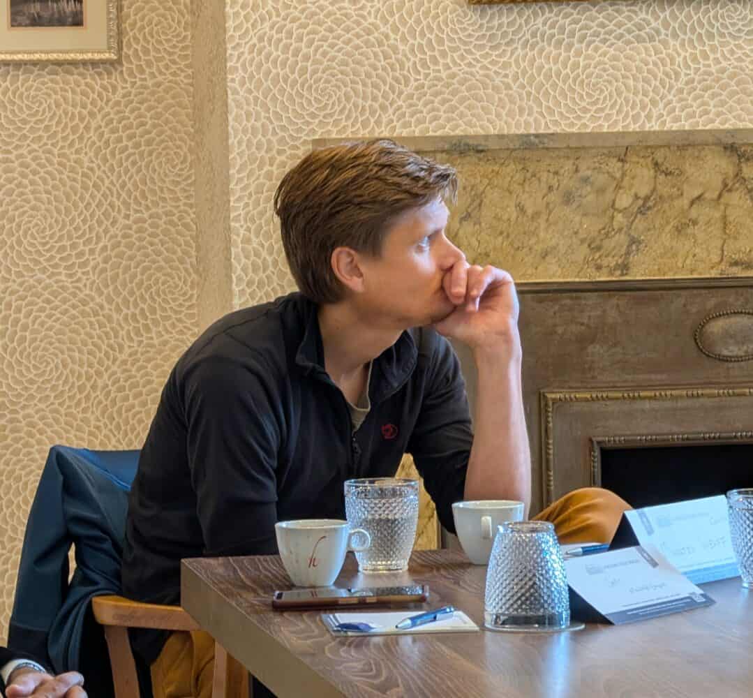 Iemand zit aan een tafel met koffiekopjes, glazen en papieren, laat zijn kin op zijn hand rusten en kijkt opzij in een contemplatieve houding.