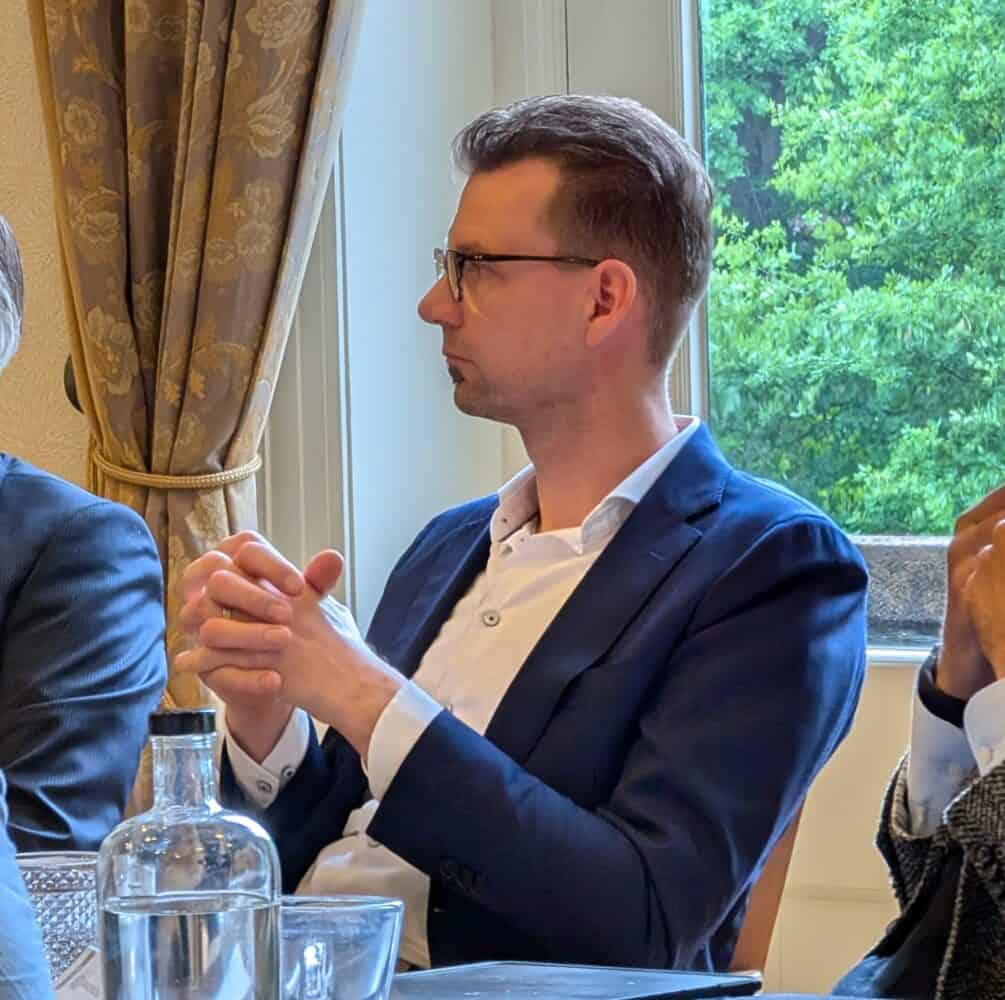 Een man met een bril en een marineblauw pak zit aan een tafel met zijn handen in elkaar geklemd en kijkt opzij. Op de achtergrond is een raam met uitzicht op groen.