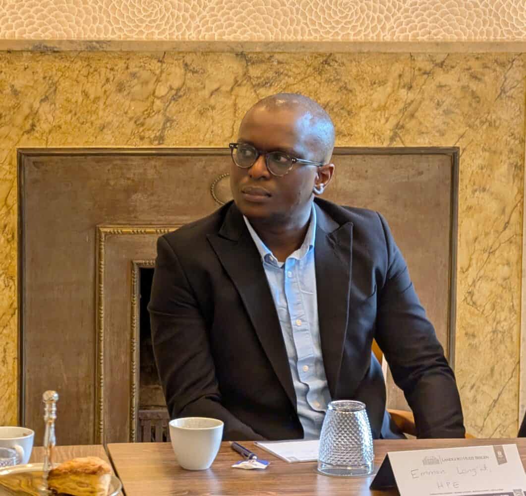 Een man in een zwart pak en blauw overhemd zit aan een tafel met kopjes, een glas en een naamkaartje voor zich, tegen een marmeren muur.