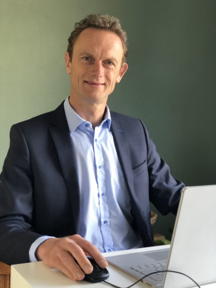 Een man in pak zit aan een bureau, gebruikt een computermuis met zijn rechterhand en lacht naar de camera. Voor hem staat een laptop open.