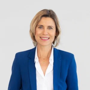 Een vrouw met kort blond haar, draagt een blauwe blazer over een wit shirt, staat tegen een effen lichte achtergrond en glimlacht naar de camera.
