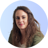 Een vrouw met lang bruin haar in een groene blazer en wit topje zit voor een lichtblauwe achtergrond en kijkt naar de camera.
