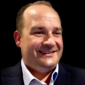 Een man met kort bruin haar, een donker colbert en lichtblauw overhemd, glimlacht tegen een effen zwarte achtergrond.