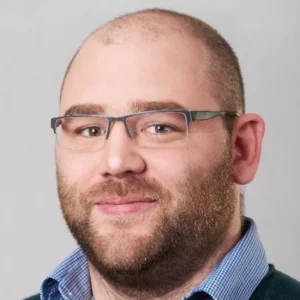 Een man met een kaal hoofd, een baard en een bril draagt een blauw overhemd en trui, kijkt met een neutrale uitdrukking naar de camera tegen een effen achtergrond.