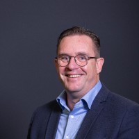Een man met kort bruin haar en een bril, draagt een blauw overhemd en donkere blazer, glimlacht voor een effen donkere achtergrond.