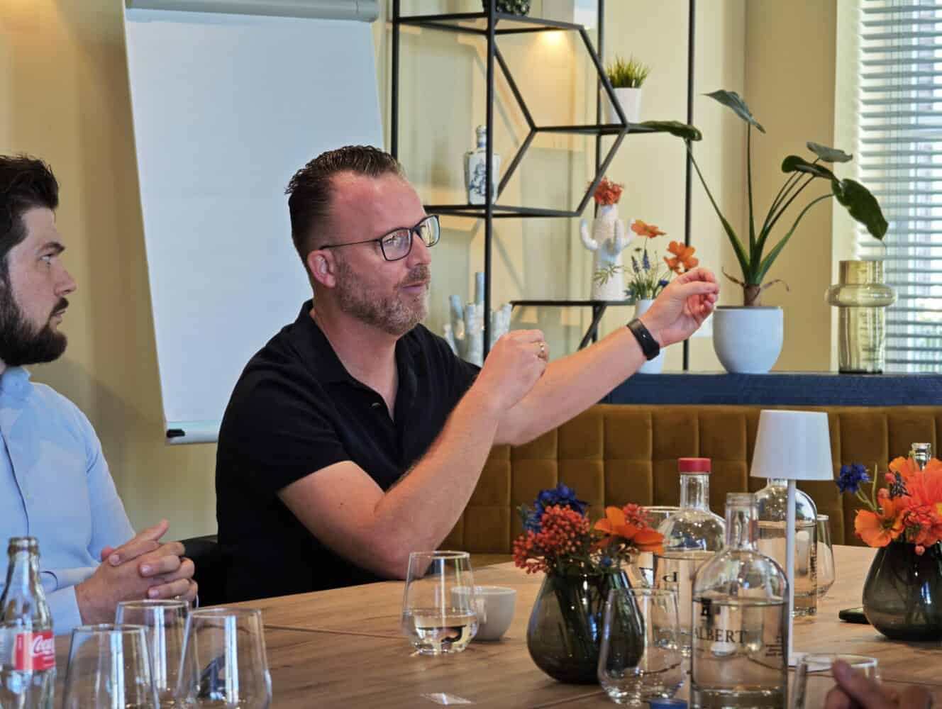 Een man met bril gebaart terwijl hij spreekt aan een vergadertafel met drankjes, bloemen en een flip-over op de achtergrond.