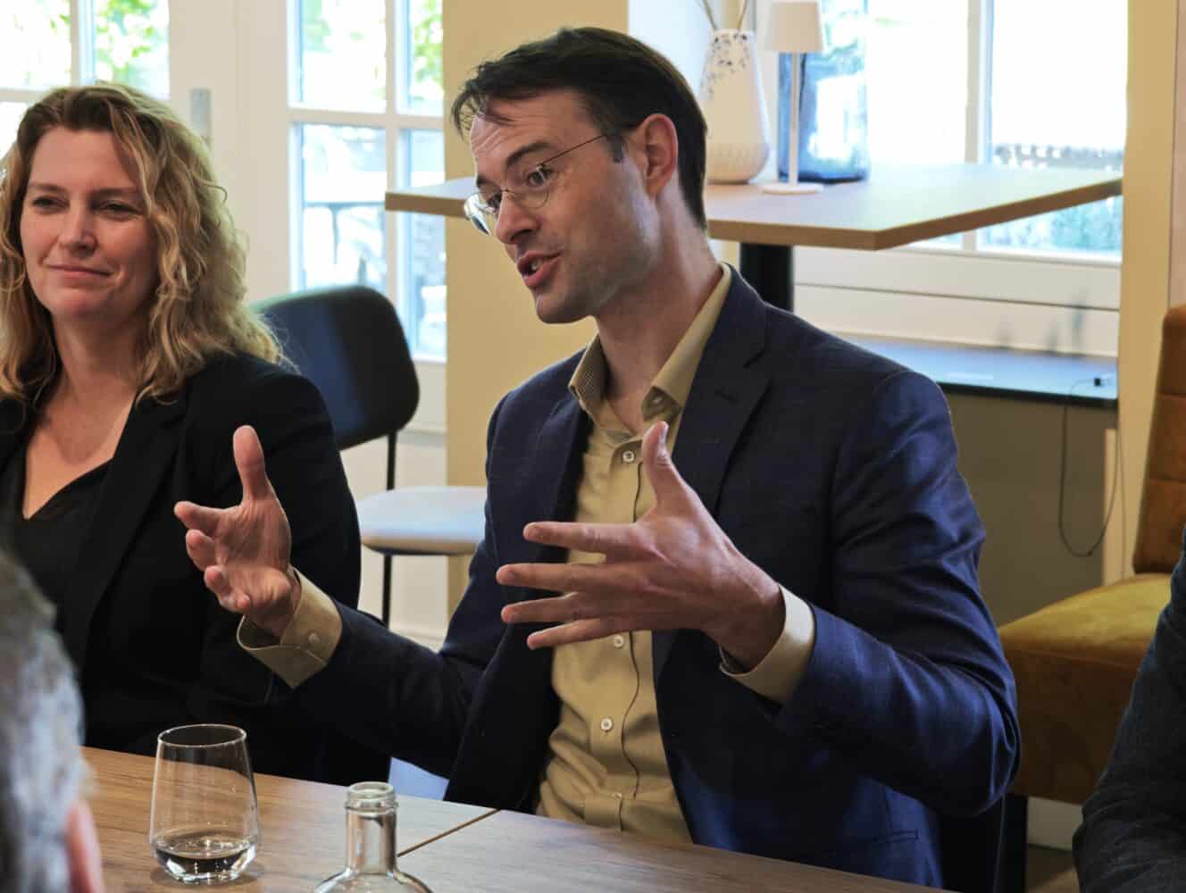 Een man in pak maakt gebaren terwijl hij spreekt aan een tafel in een vergadering, met een vrouw naast hem en een glas water voor hen.