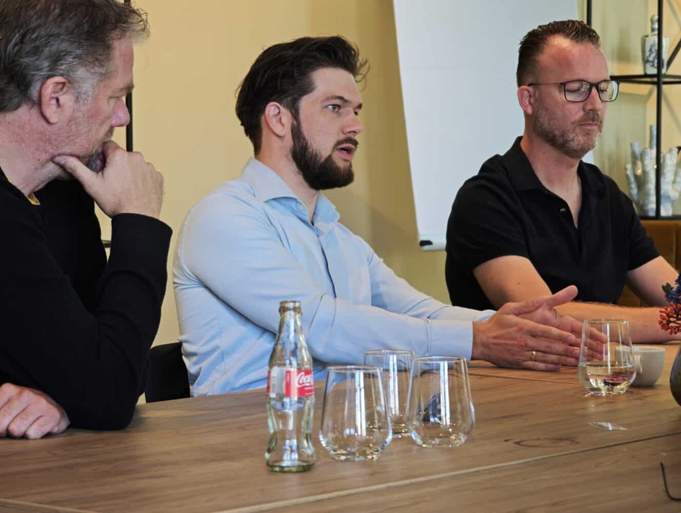 Drie mannen zitten aan een tafel te discussiëren; er staan lege glazen en een Coca-Cola fles op tafel. Eén man gebaart terwijl hij spreekt, de anderen luisteren aandachtig.