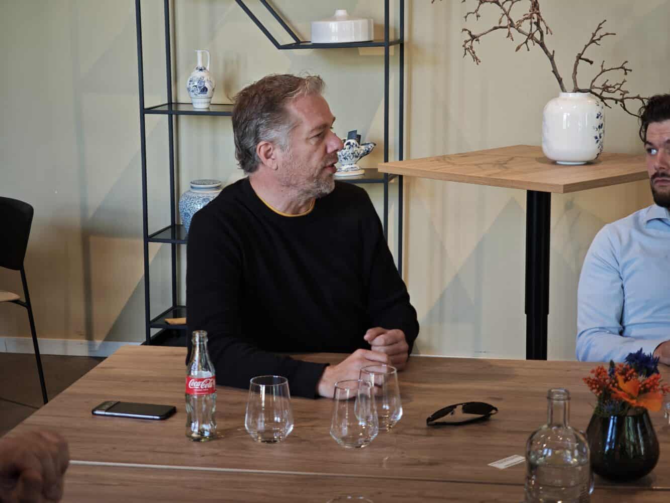 Een man in een zwarte trui zit aan een houten tafel met glaswerk, een Coca-Cola fles en bloemen en praat met een andere man in een lichtblauw shirt.