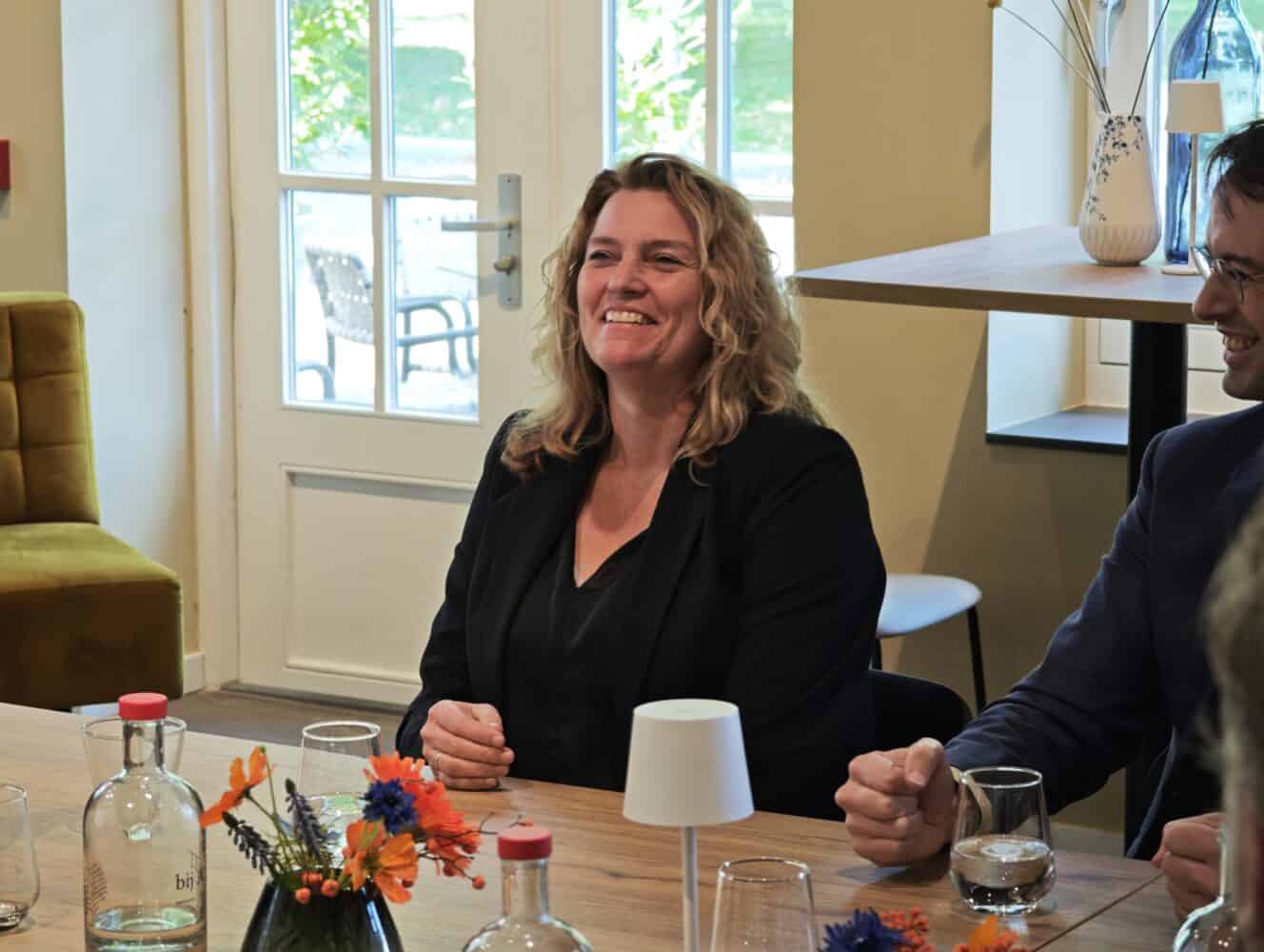 Een vrouw met golvend blond haar en een zwarte blazer zit aan een tafel met drankjes en bloemen en glimlacht tijdens een vergadering in een goed verlichte kamer.