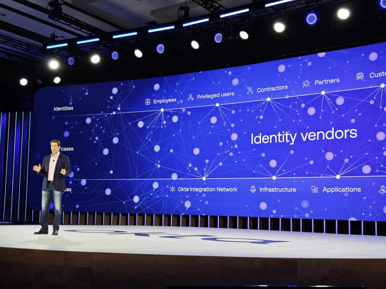 Een man staat op het podium te presenteren voor een groot scherm met daarop "Identity vendors" met aanverwante termen als Okta en network graphics.
