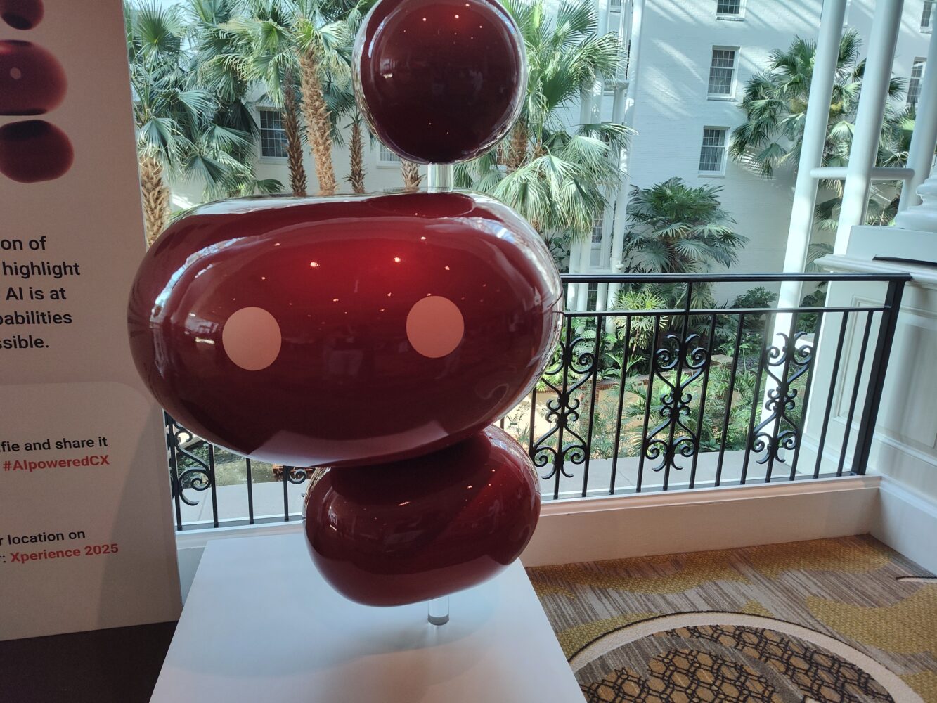 Een glanzende, abstracte Genesys sculptuur met drie gestapelde kastanjebruine bollen en witte ronde accenten staat binnen bij een reling en palmbomen.