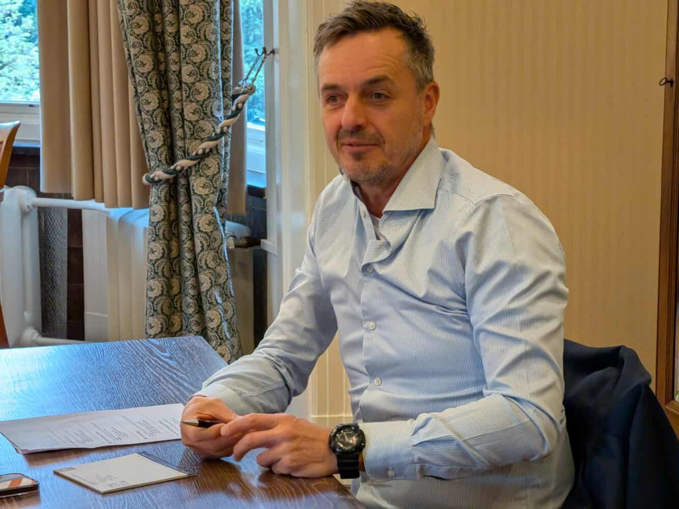 Een man in een lichtblauw overhemd zit aan een houten tafel met documenten en een pen en kijkt een beetje opzij.
