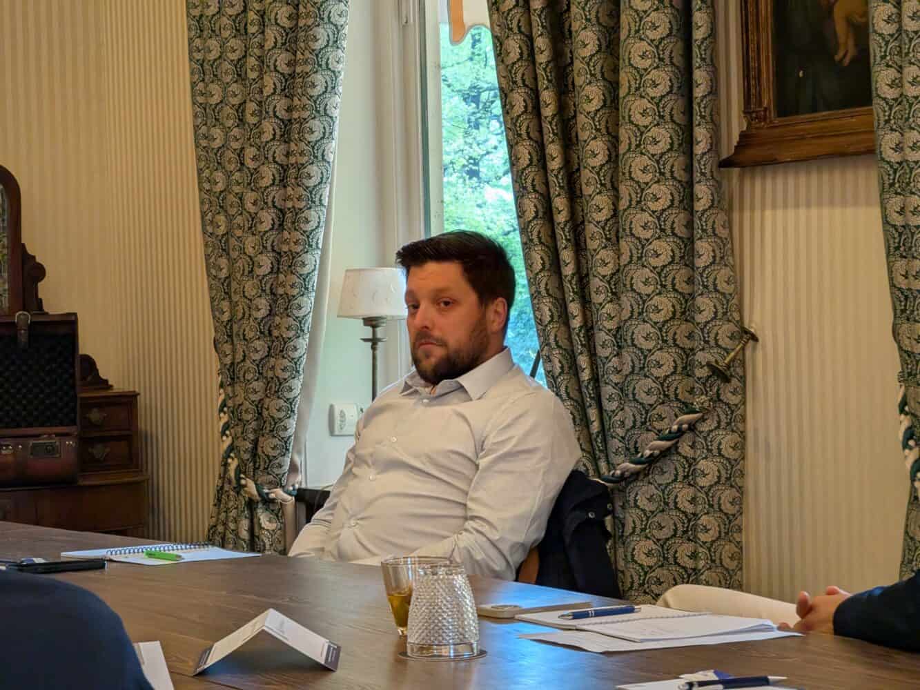 Een man in een wit overhemd zit aan een tafel met papieren, een glas water en een notitieblok voor zich, in een kamer met gedessineerde gordijnen en een raam.