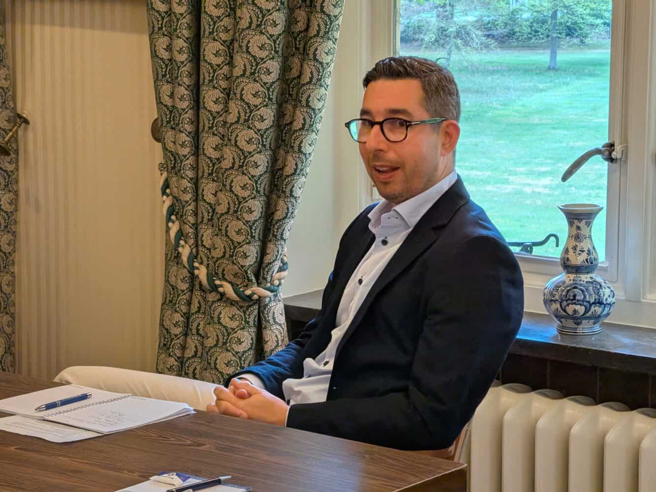 Een man met een bril en een donkere blazer zit aan een tafel met papieren en een pen te praten. Achter hem is een raam met gedessineerde gordijnen en een decoratieve vaas.