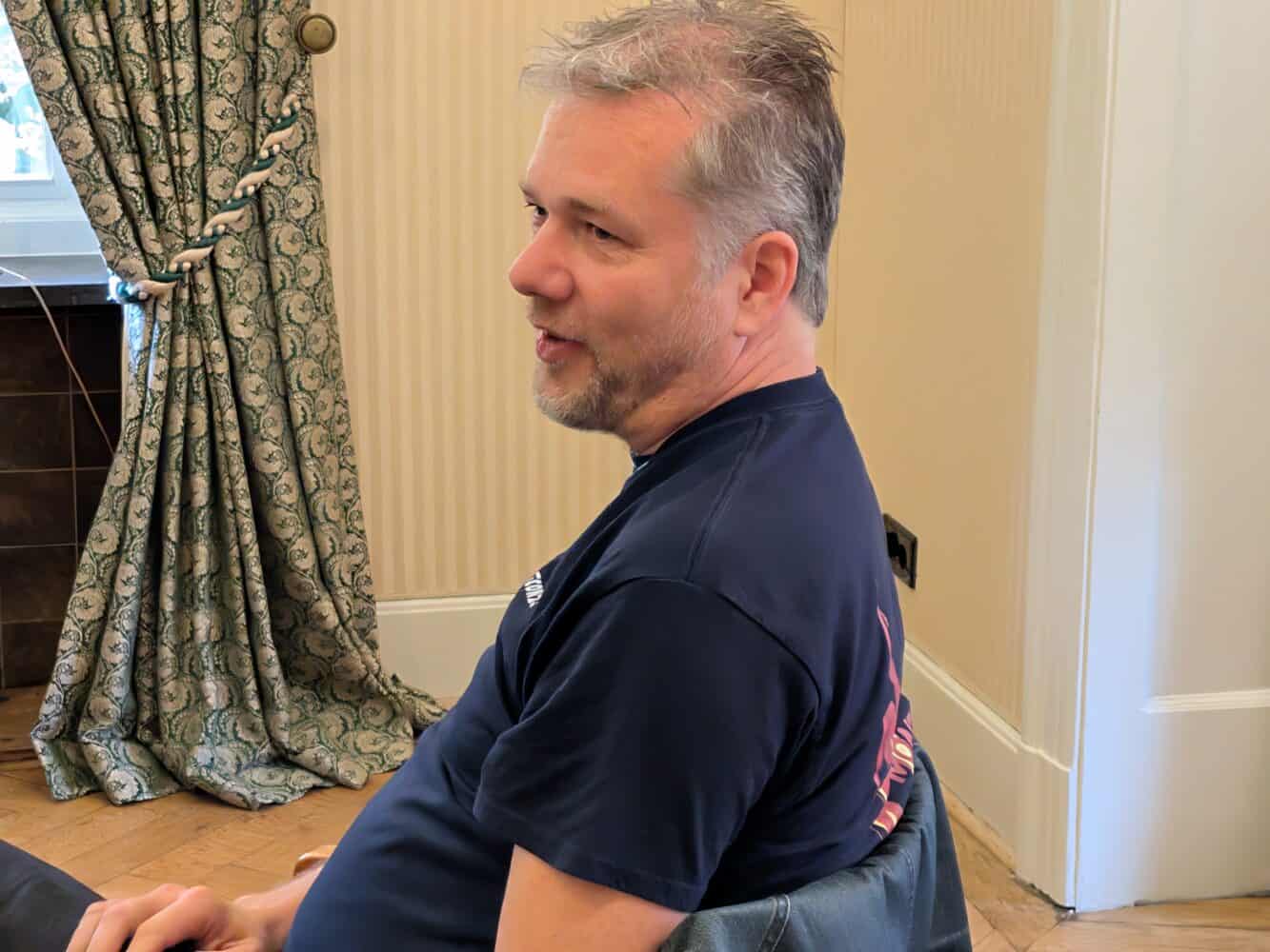 Een man met kort grijs haar en een baard zit en praat in een kamer met gedessineerde gordijnen en crèmekleurige muren.
