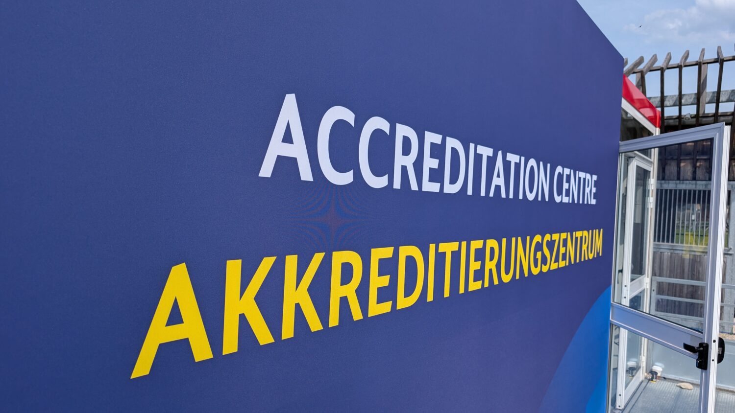 Groot blauw bord met "ACCREDITATION CENTRE" in het Engels en "AKKREDITIERUNGSZENTRUM" in het Duits bij een open glazen deur.