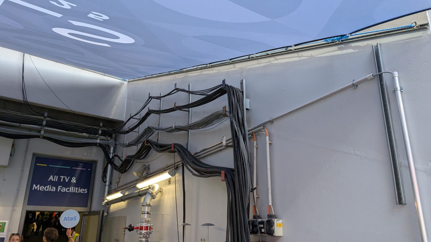 Dikke zwarte kabels zijn gebundeld en lopen langs de muren en het plafond bij een deur met het label "Alle TV & Media faciliteiten" in een industriële omgeving.