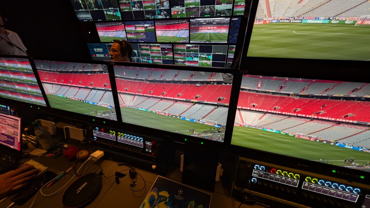 Meerdere monitoren in een controlekamer tonen live beelden van een voetbalstadion met lege rode en grijze stoelen voorafgaand aan een evenement.