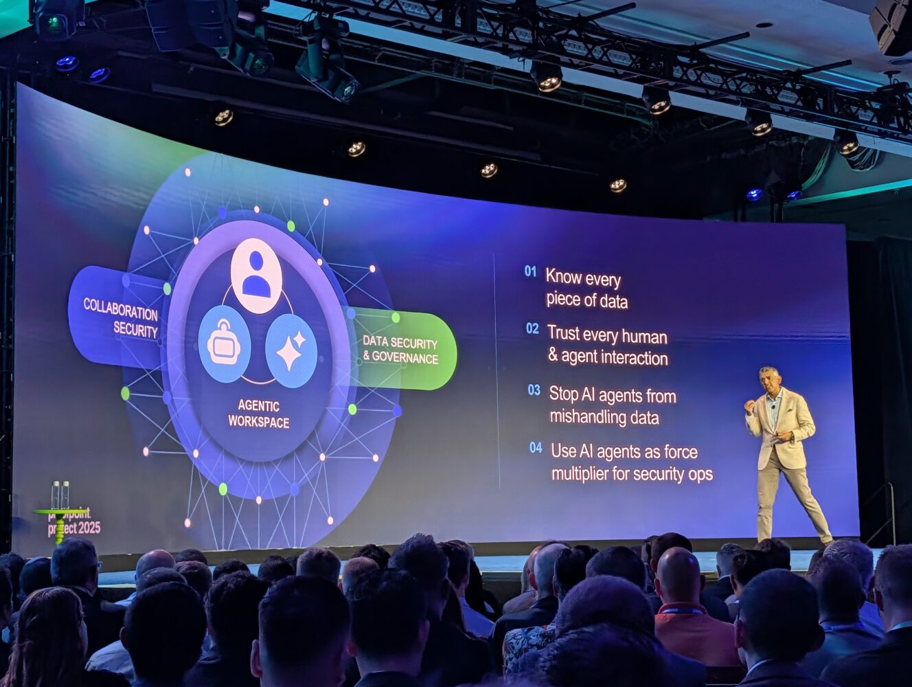 Een spreker presenteert op een podium met een groot scherm waarop informatie wordt weergegeven over samenwerkingsbeveiliging, gegevensbeveiliging, governance en AI-agentstrategieën, waaronder Proofpoint-oplossingen, aan een publiek op een conferentie.