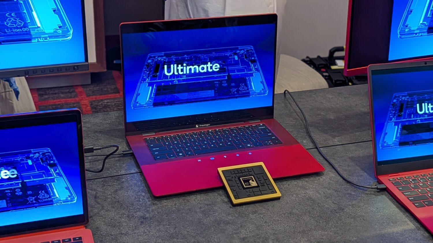 Een rode laptop op een tafel toont het woord "Ultimate" op het scherm, met twee andere laptops in de buurt en een vierkant apparaat voor de centrale laptop.