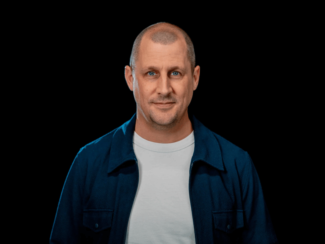 Een man van middelbare leeftijd met kort haar en blauwe ogen, die een wit T-shirt en een blauw jasje draagt, staat tegen een effen zwarte achtergrond.
