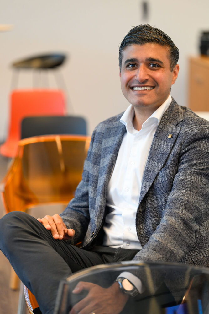Een man in een geruite blazer en wit overhemd zit in kleermakerszit op een stoel en lacht naar de camera. Op de achtergrond staan meer stoelen en wazig kantoormeubilair.
