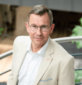 Een man met een bril en een lichtgekleurde blazer staat binnen en kijkt met een neutrale uitdrukking naar de camera.