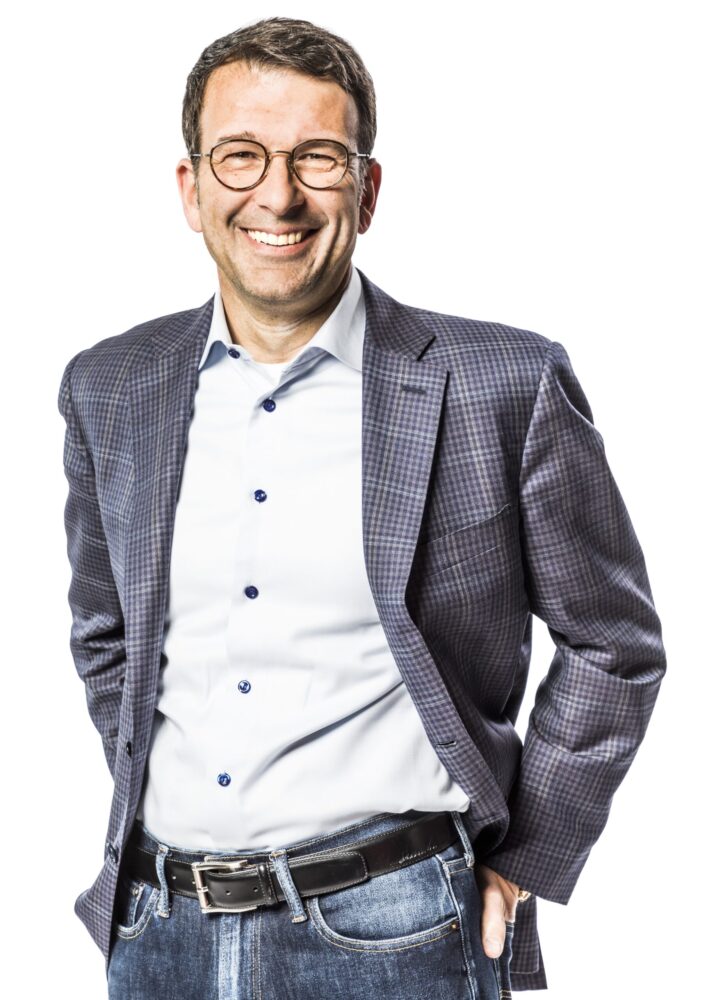 Man met bril, geruite blazer, wit overhemd en spijkerbroek, staand en lachend met handen in zakken tegen een witte achtergrond.