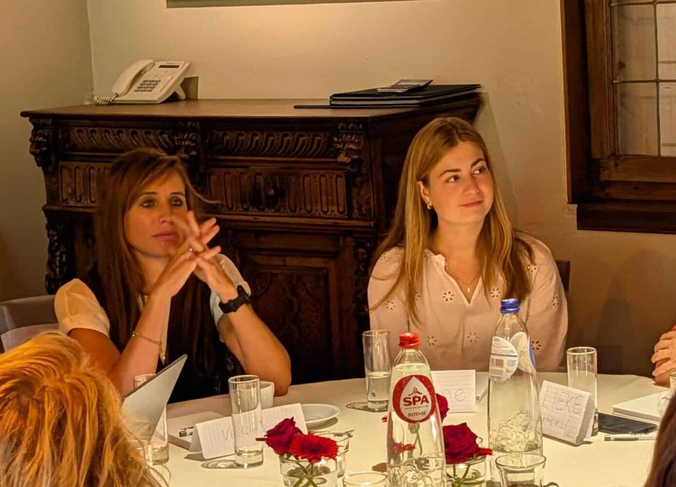 PXL_20250916_084337628 Een groep mensen zit rond een ronde tafel in een goed verlichte kamer en is in gesprek. Er liggen papieren, glazen en een fles water op tafel.