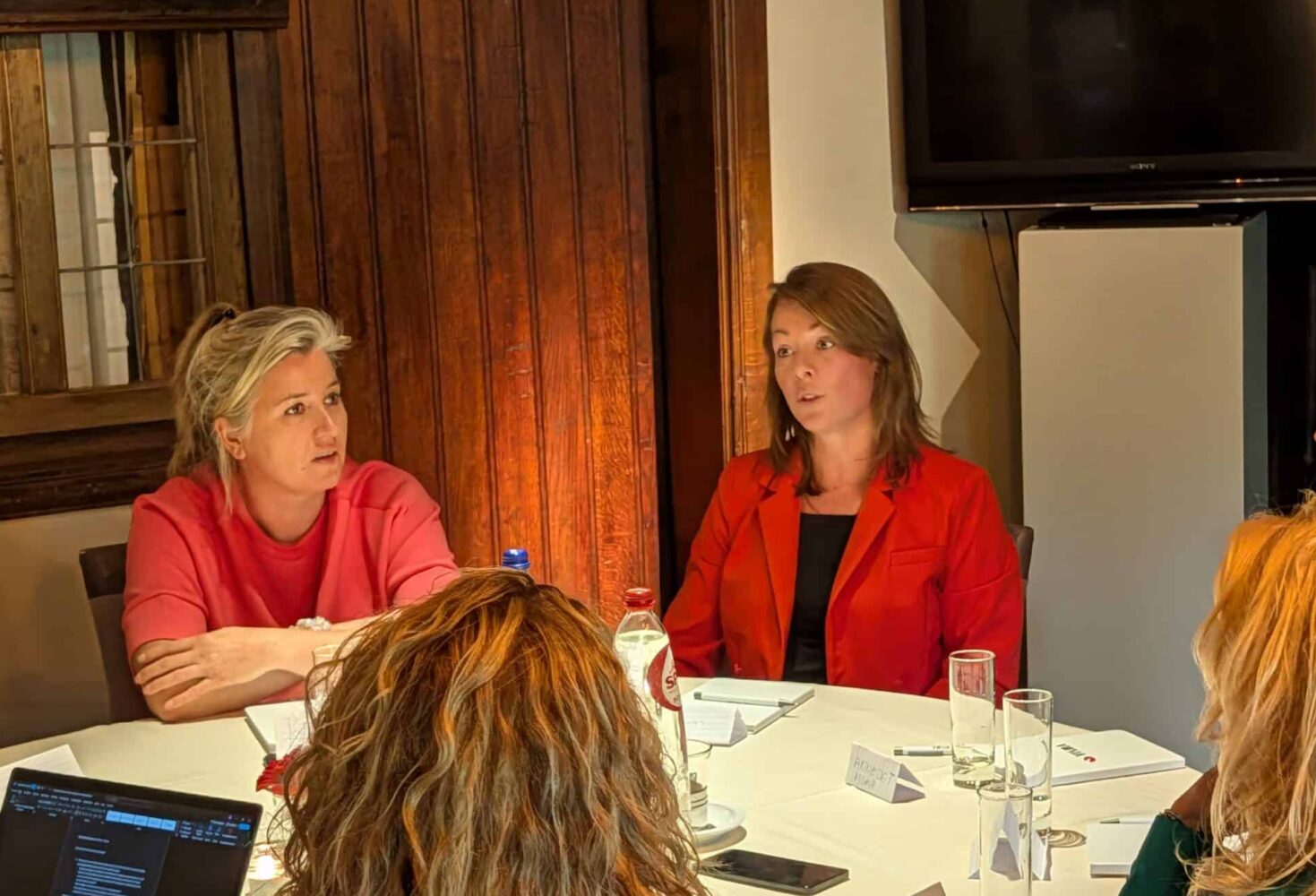 PXL_20250916_084349839.TS-000 Zes vrouwen zitten rond een tafel en discussiëren met documenten, laptops en glazen op tafel in een kamer met houten panelen en een tv aan de muur.