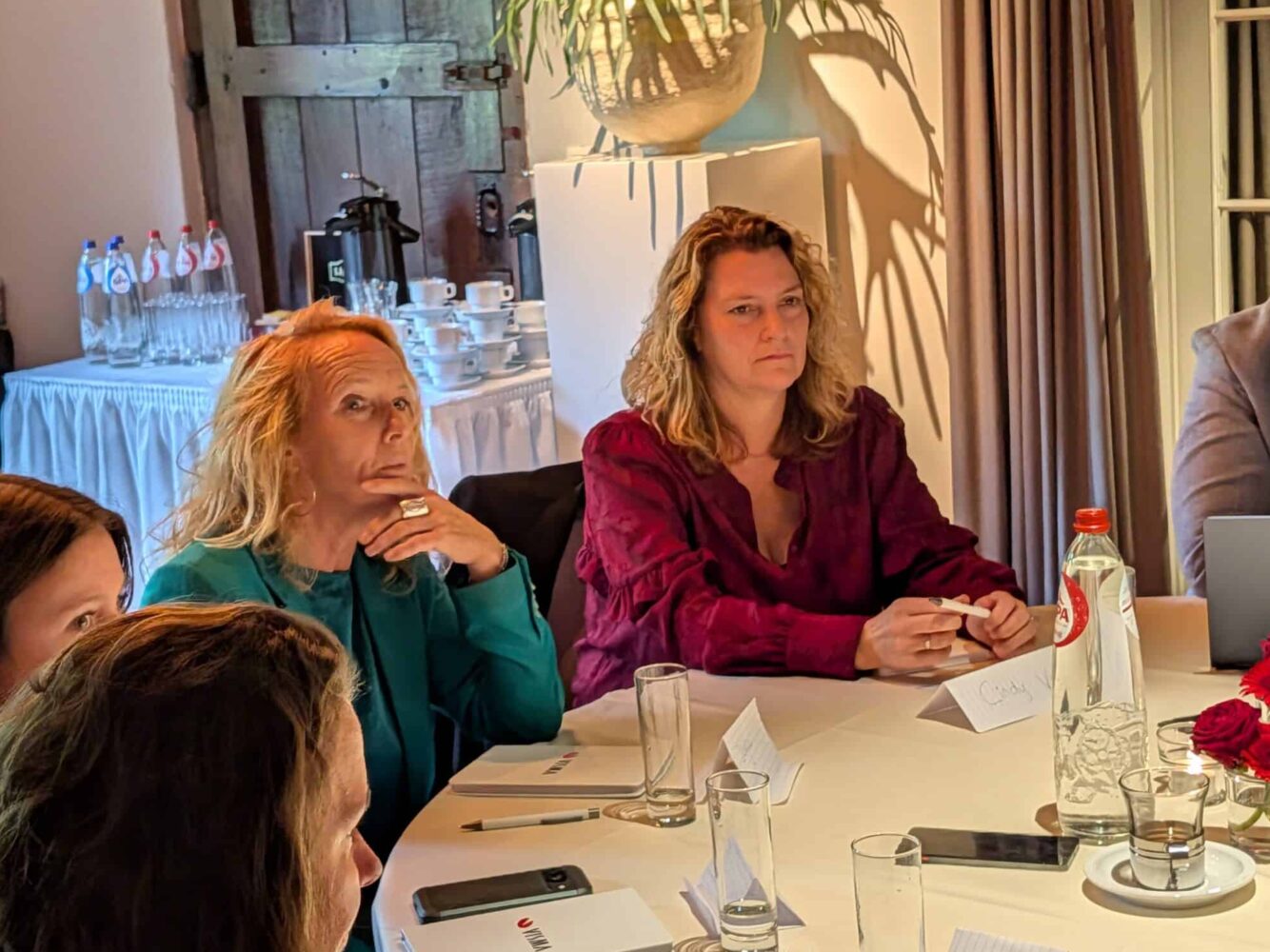 PXL_20250916_084407495 Vijf mensen zitten rond een ronde tafel met blocnotes, waterflessen en glazen, bezig met een vergadering in een goed verlichte kamer.
