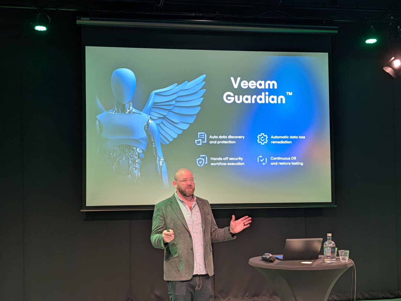 PXL_20250918_091427255.MP Een man staat en gebaart terwijl hij voor een scherm staat met de titel "Veeam Guardian" en een afbeelding van een gevleugelde mensachtige figuur en beveiligingsfuncties opgesomd.