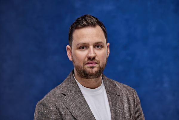Man met kort bruin haar en baard draagt een grijs geruite blazer over een wit t-shirt, poserend voor een effen blauwe achtergrond.