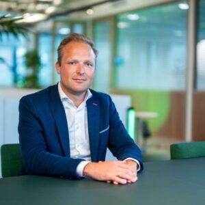Een man in een blauw pak zit aan een tafel in een modern kantoor met glazen muren en groene accenten, en kijkt recht in de camera.