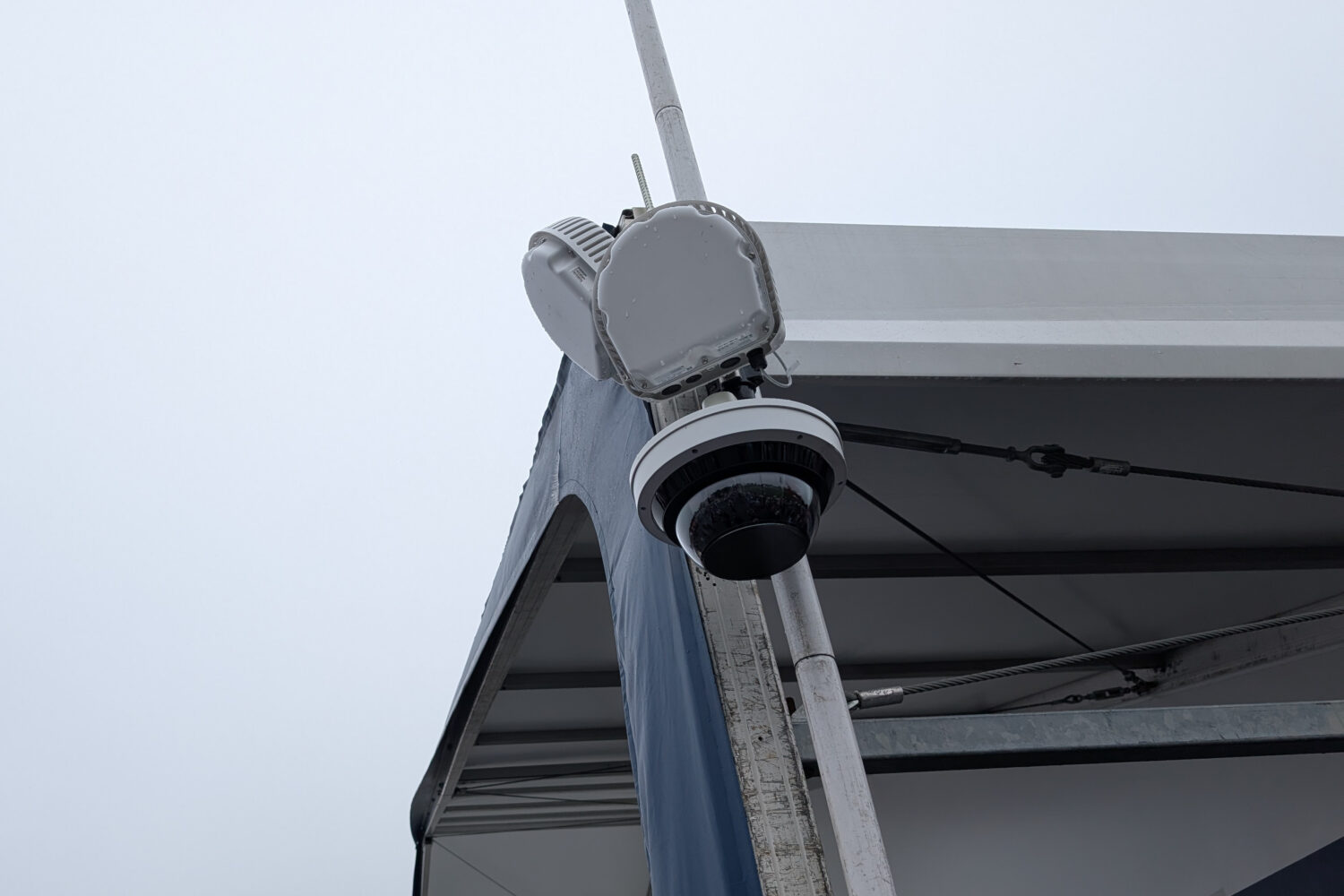 AP_Camera_Ryder_Cup Een beveiligingscamera en communicatie-apparaat gemonteerd op een metalen paal naast een structuur met een blauwe en witte luifel, tegen een bewolkte lucht.