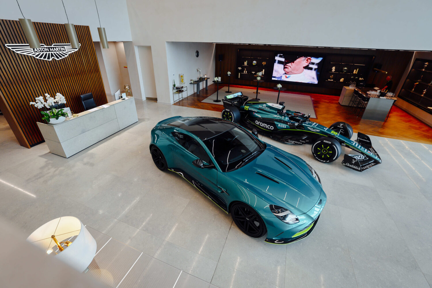 Een groenblauwe Aston Martin sportwagen en een Formule 1-racewagen staan tentoongesteld in een moderne showroom met een receptie en een groot scherm aan de muur.
