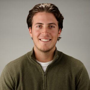 Een jonge man met bruin haar, die een groene trui met ritssluiting over een wit overhemd draagt, glimlacht naar de camera tegen een effen grijze achtergrond.