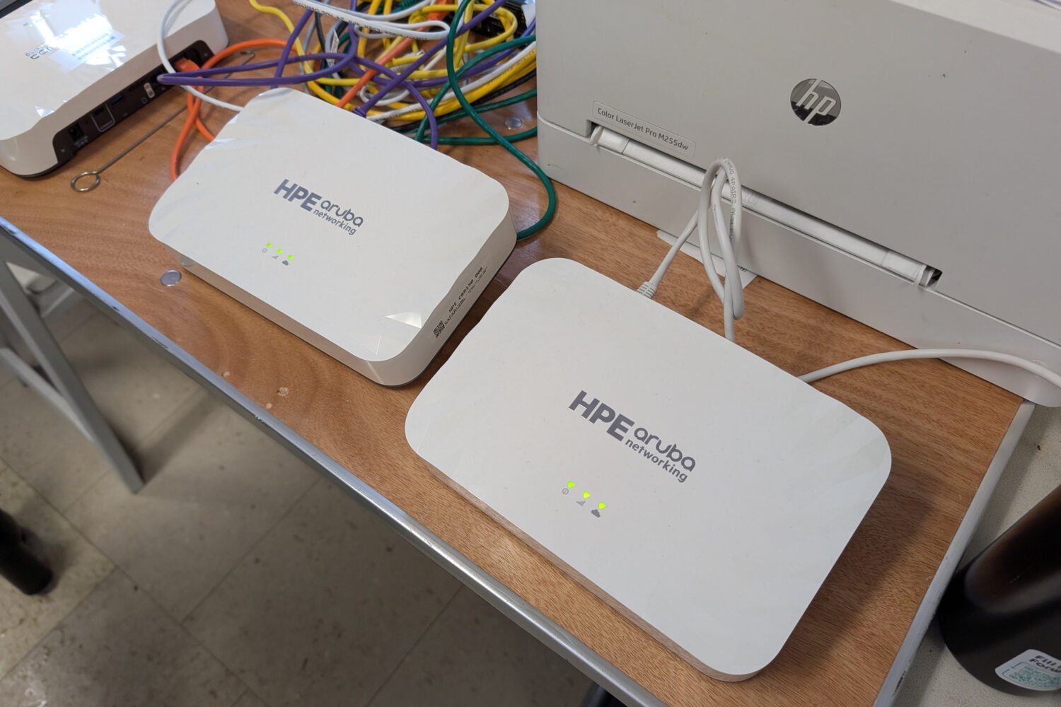 HPE_Aruba_BR150 Twee HPE Aruba netwerkapparaten met indicatielampjes staan op een bureau, in de buurt van een printer en verwarde netwerkkabels.
