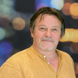 Een man van middelbare leeftijd met kort bruin haar en een lichte baard draagt een mosterdgeel overhemd en staat voor een wazige achtergrond met kleurrijke lichten.