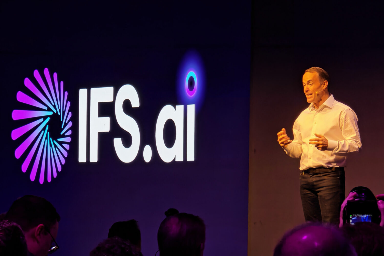 Een man in een wit shirt spreekt op het podium naast een groot 