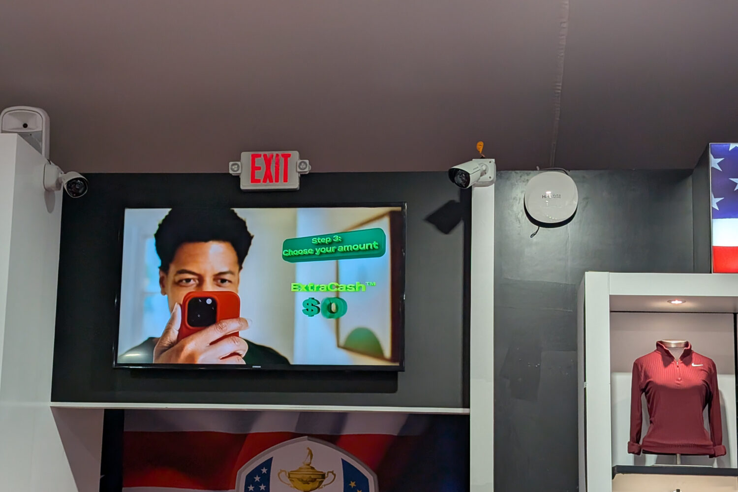 Op een tv-scherm is een persoon te zien die een telefoon vasthoudt met de tekst "Stap 2: kies uw bedrag. ExtraCash™ $0", boven een vitrine met een kastanjebruin overhemd met ritssluiting.