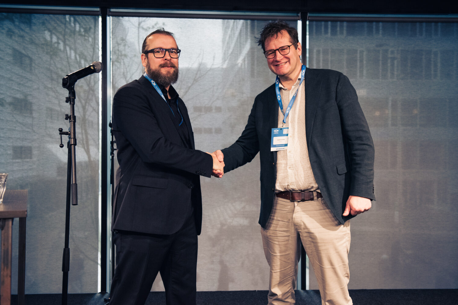 Twee mannen in zakelijke kleding schudden elkaar de hand en glimlachen voor een groot raam, met links een microfoonstandaard van KPN.